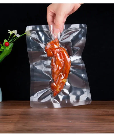 Hand holding a vacuum-sealed package of a roasted chicken leg on a wooden surface with a dark background.