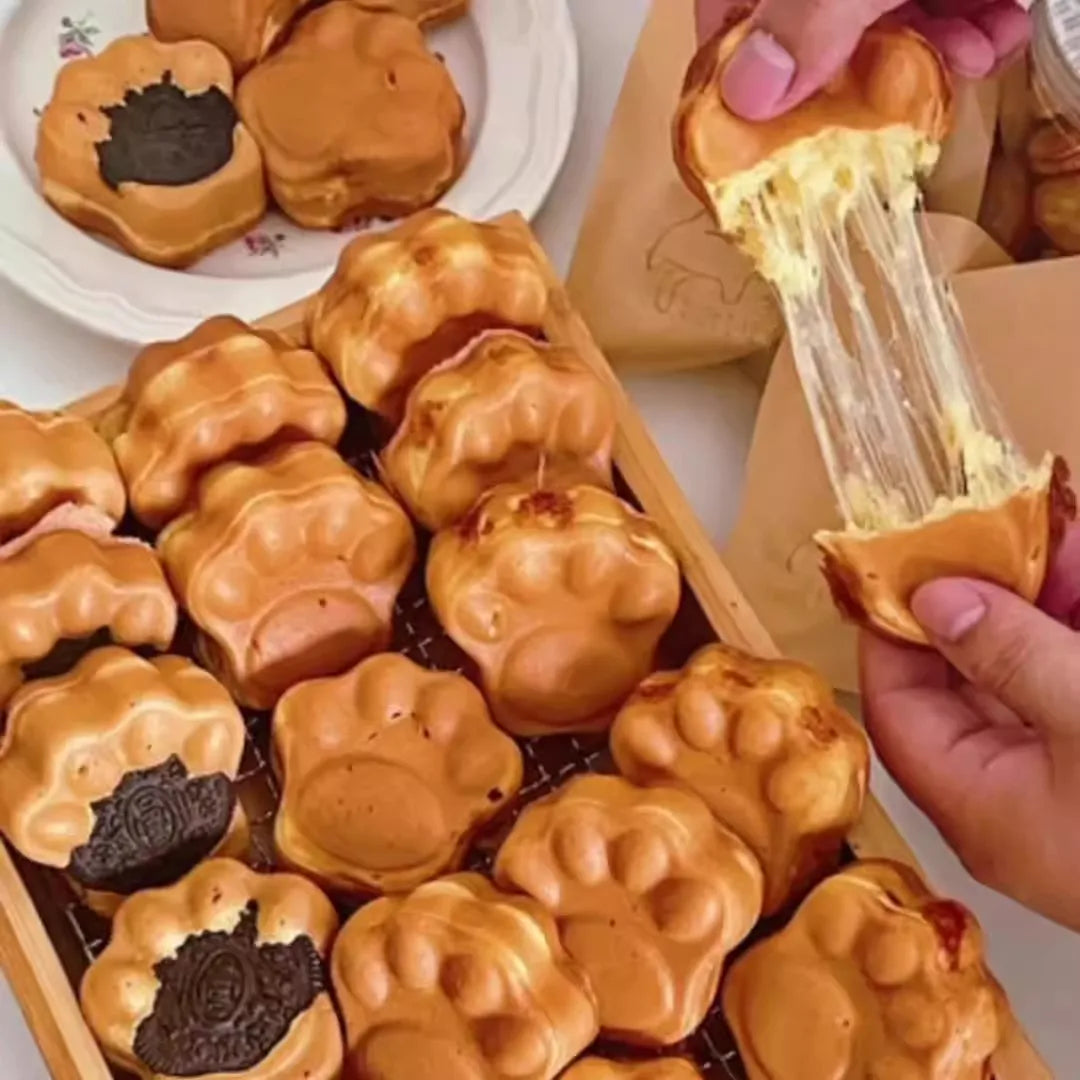 Golden-brown paw-shaped waffle pastries arranged on a wooden tray, with a hand pulling apart one to show a stretchy, creamy filling. Some pastries are topped with dark chocolate cookie pieces on a white plate in the background.